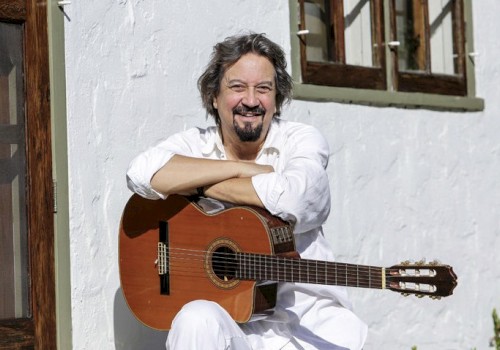 A smiling man in white clothes sits outside with an acoustic guitar resting on his lap, posing beside a white wall and window.