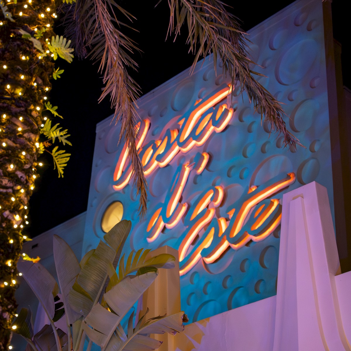 A neon sign is displayed against a textured blue wall with tropical plants and palm branches lit by decorative lights.
