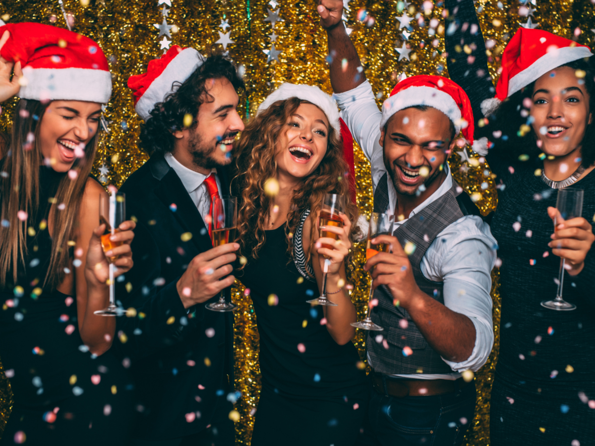 A group of people wearing Santa hats, smiling, and holding drinks is celebrating against a festive, glittery backdrop with confetti falling.