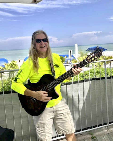 A person with long hair and sunglasses plays a guitar on a sunny beachside deck, wearing a bright yellow shirt, next to the ocean.