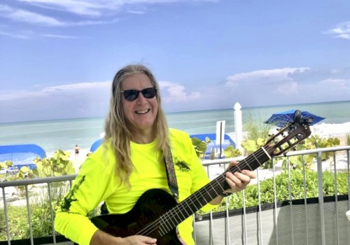 A person with long hair and sunglasses plays a guitar on a sunny beachside deck, wearing a bright yellow shirt, next to the ocean.