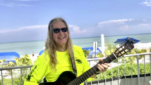 A person with long hair and sunglasses plays a guitar on a sunny beachside deck, wearing a bright yellow shirt, next to the ocean.