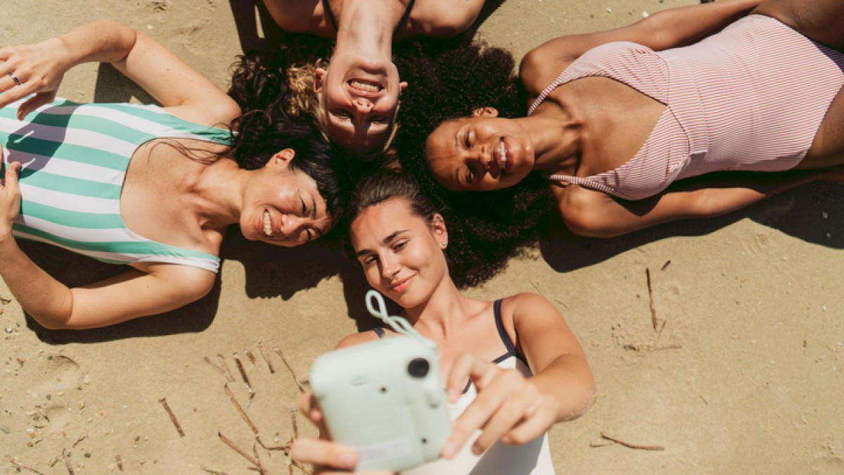 Four women are lying on the sand taking a selfie together on the beach.