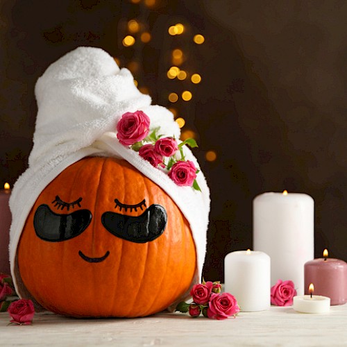 A cute pumpkin with a towel turban, fake eyelashes, and pink roses, surrounded by candles, creating a cozy and festive fall scene.