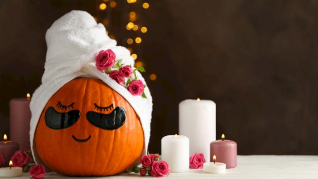 A cute pumpkin with a towel turban, fake eyelashes, and pink roses, surrounded by candles, creating a cozy and festive fall scene.