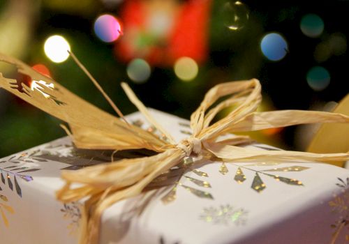 The image shows a beautifully wrapped gift adorned with a gold bow and decorative paper, with festive lights blurred in the background.