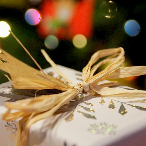 The image shows a beautifully wrapped gift adorned with a gold bow and decorative paper, with festive lights blurred in the background.