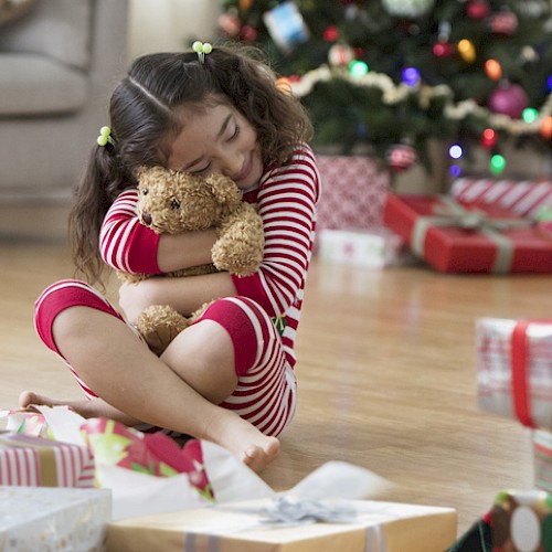 A young girl in holiday pajamas hugs a teddy bear next to a decorated Christmas tree surrounded by wrapped presents.