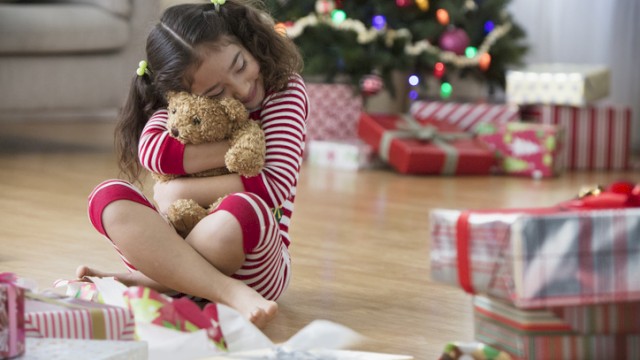 A young girl in holiday pajamas hugs a teddy bear next to a decorated Christmas tree surrounded by wrapped presents.