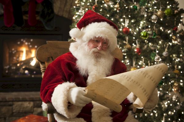 A man dressed as Santa Claus reading a long list while sitting in front of a fireplace and a decorated Christmas tree.
