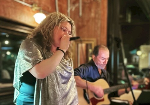 A woman passionately sings into a microphone while a man guitarist plays on, both performing in a cozy, intimate setting.