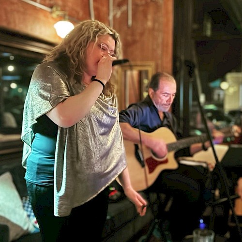 A woman passionately sings into a microphone while a man guitarist plays on, both performing in a cozy, intimate setting.