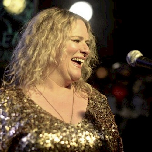 A woman with curly blonde hair laughing on stage, wearing a gold sequined top, with a microphone in front of her at a lively event.