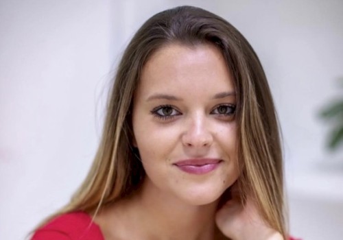 A young woman with long hair in a red top, smiling softly, sitting indoors with a blurred background.