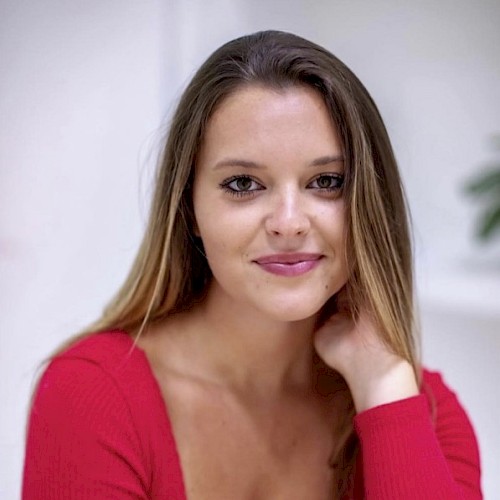 A young woman with long hair in a red top, smiling softly, sitting indoors with a blurred background.