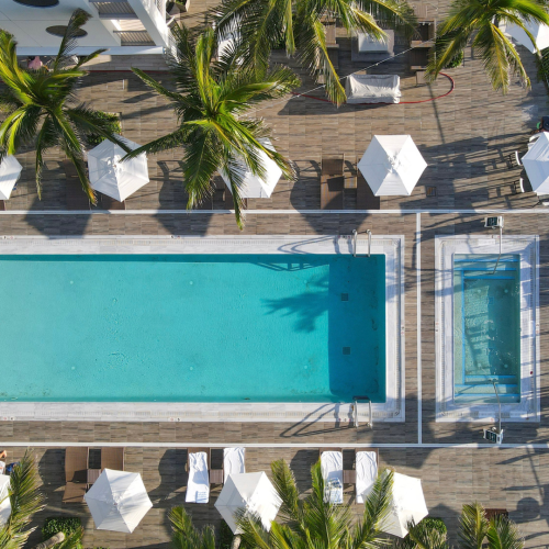 An aerial view of a resort pool area with chairs, umbrellas, palm trees, and a surrounding wooden deck.