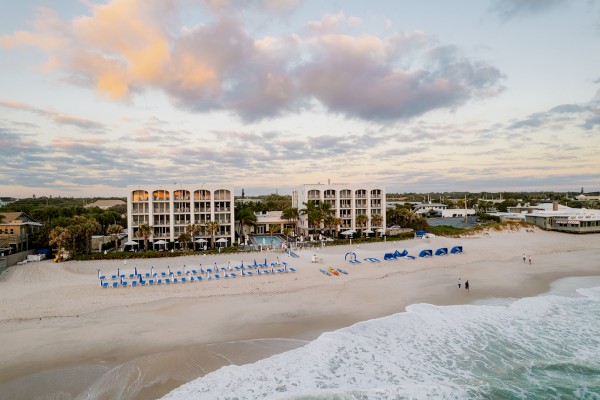 A beachside resort with white buildings, palm trees, lounge chairs, umbrellas, paddleboards, cabanas, and people enjoying the sand and ocean.