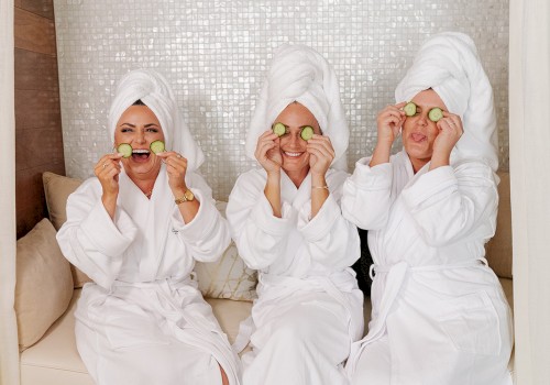 Three people in white bathrobes with towels on their heads, sitting on a sofa, placing cucumber slices over their eyes, enjoying a spa day.