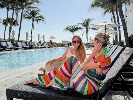 Two women are relaxing on lounge chairs by a pool with palm trees, sunny sky, and umbrellas visible in the background.