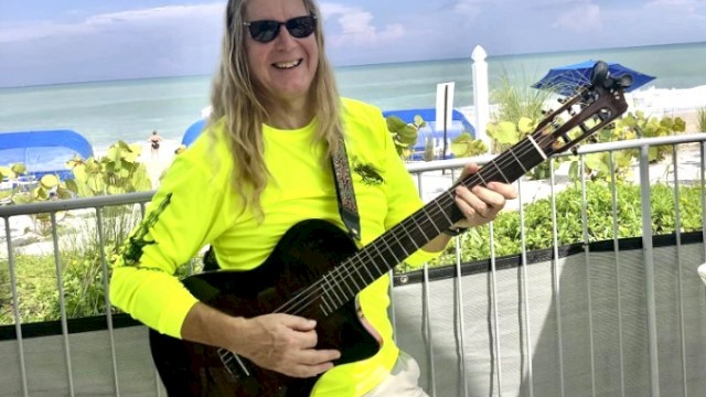 A person in sunglasses and a neon shirt plays guitar by the beach, with blue umbrellas and ocean in the background.