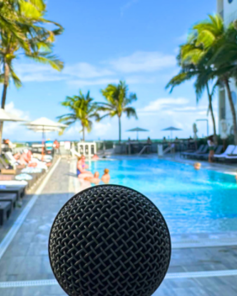 A microphone is in the foreground with a vibrant pool scene behind; palm trees, lounge chairs, and people enjoying the sunny weather.