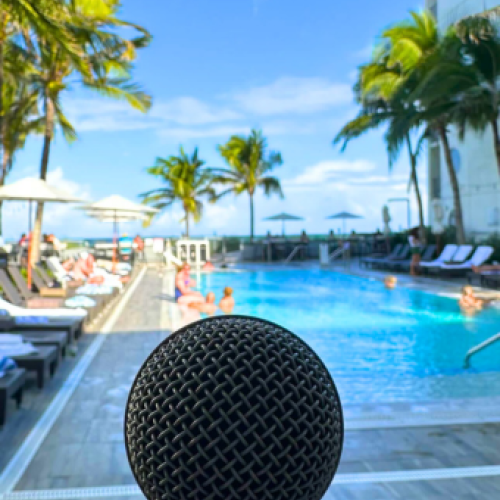 A microphone is in the foreground with a vibrant pool scene behind; palm trees, lounge chairs, and people enjoying the sunny weather.