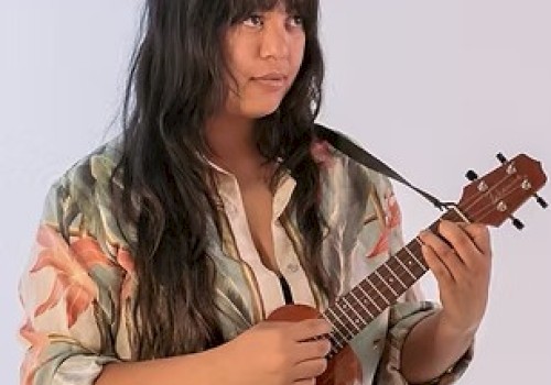 A woman sits and strums a small wooden ukulele, wearing a floral shirt with long dark hair, focused on playing.