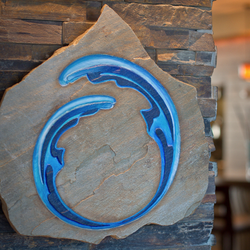 A stylized, blue wave design is carved on a rock surface, with a blurred bar area in the background.