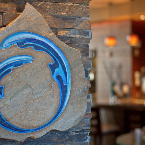 A stylized, blue wave design is carved on a rock surface, with a blurred bar area in the background.