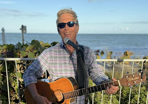 A man wearing sunglasses plays a guitar and sings into a microphone by the seaside, under a clear blue sky.