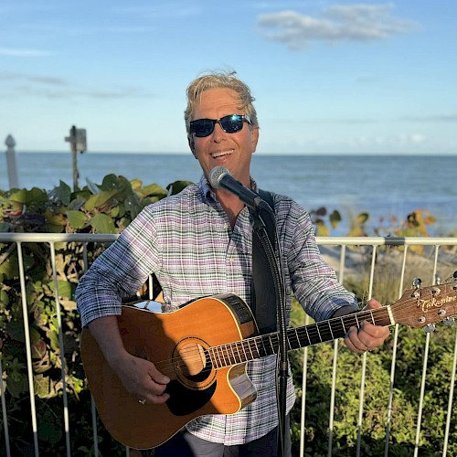 A man wearing sunglasses plays a guitar and sings into a microphone by the seaside, under a clear blue sky.