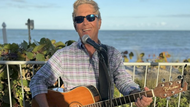 A man wearing sunglasses plays a guitar and sings into a microphone by the seaside, under a clear blue sky.
