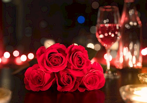A romantic setting with red roses, a glass of wine, a bottle, and candles on a dimly lit table.