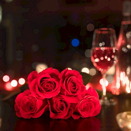 A romantic setting with red roses, a glass of wine, a bottle, and candles on a dimly lit table.