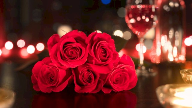 A romantic setting with red roses, a glass of wine, a bottle, and candles on a dimly lit table.