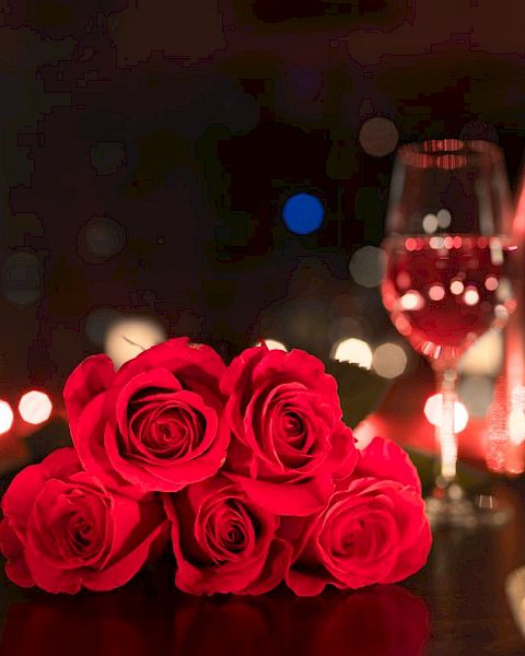 A romantic setting with red roses, a glass of wine, a bottle, and candles on a dimly lit table.