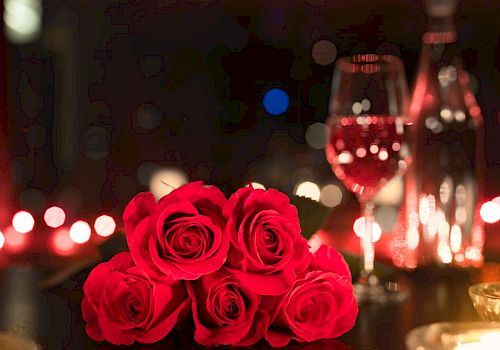 A romantic setting with red roses, a glass of wine, a bottle, and candles on a dimly lit table.