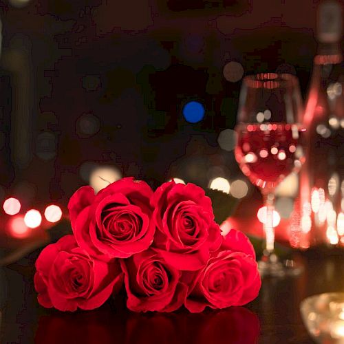 A romantic setting with red roses, a glass of wine, a bottle, and candles on a dimly lit table.