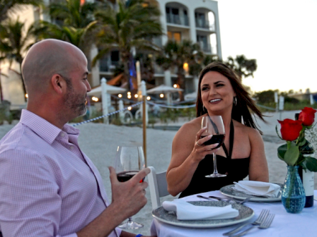 A man and woman enjoy wine and conversation at an outdoor dinner with a seaside hotel in the background.