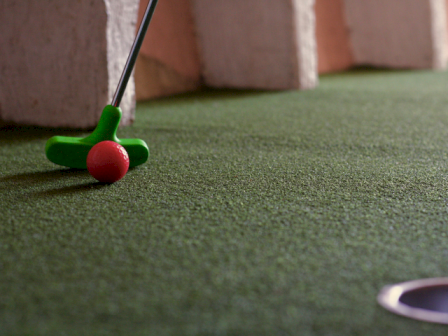 A miniature golf putting green with a practice golf club and a pink golf ball near a hole, likely indoors or in a miniature course area.