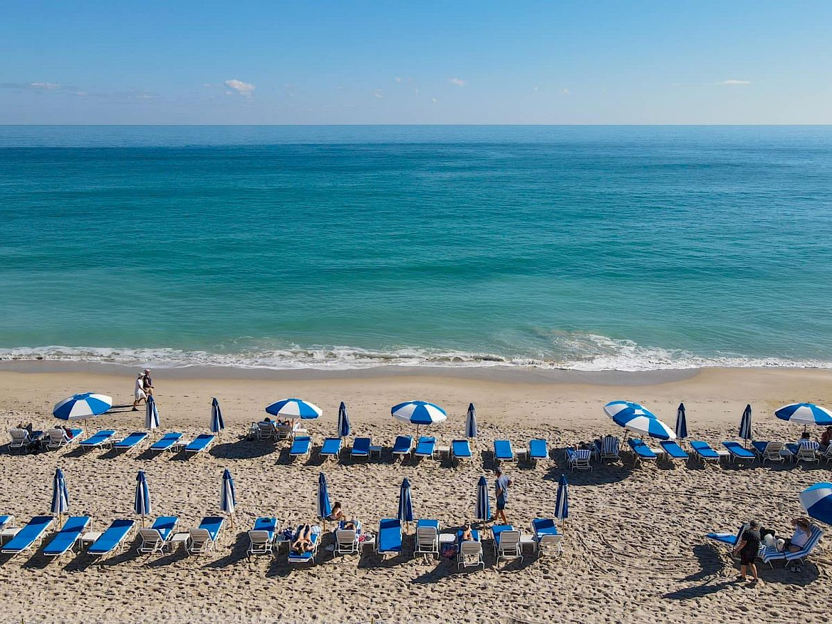 A tranquil beach with blue and white umbrellas, lounge chairs, and a few people relaxing near the clear blue ocean.