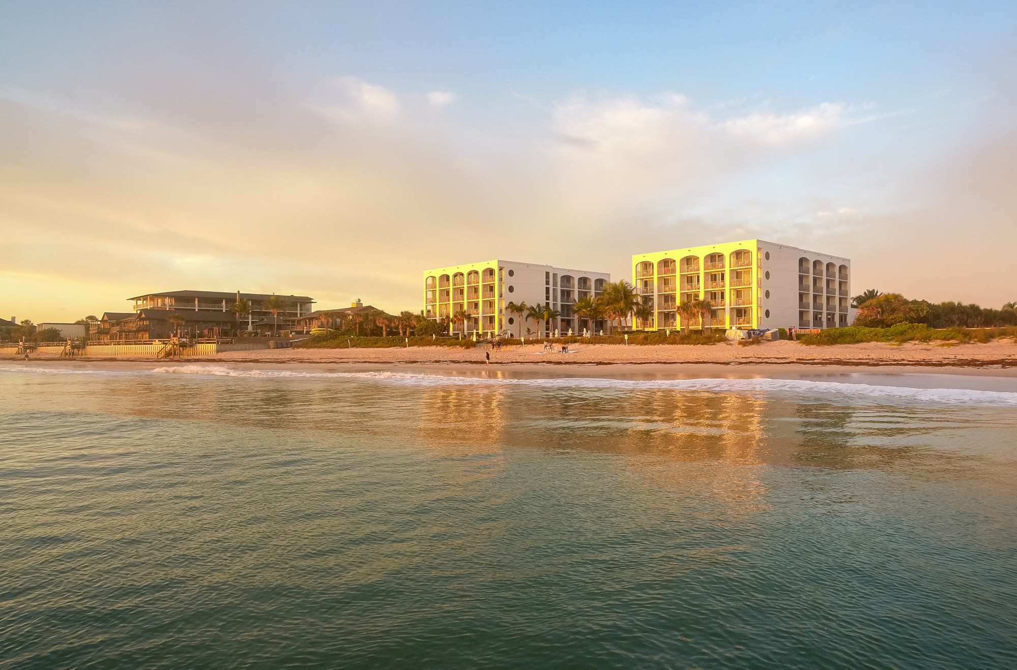 A serene beach scene with calm water, sandy shore, and two modern buildings in the background under a partly cloudy sky bathed in soft light.