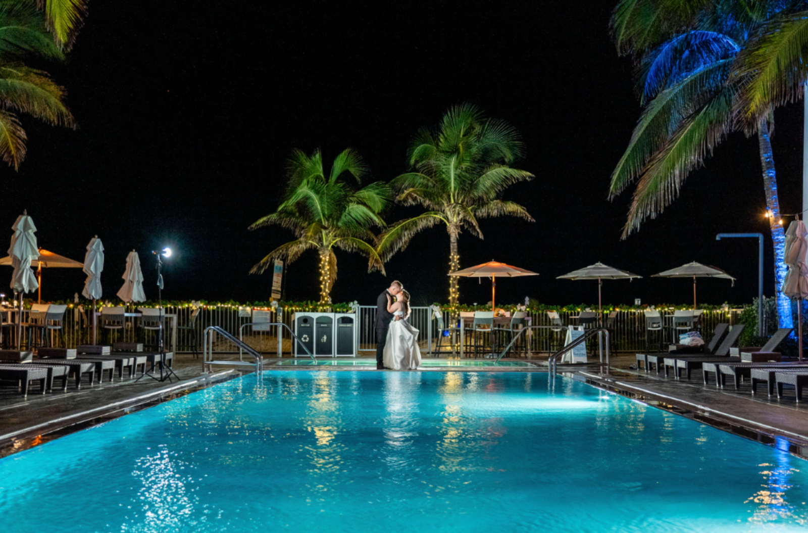 A couple stands by a pool at night, surrounded by palm trees and decorated lights, creating a romantic, serene atmosphere.