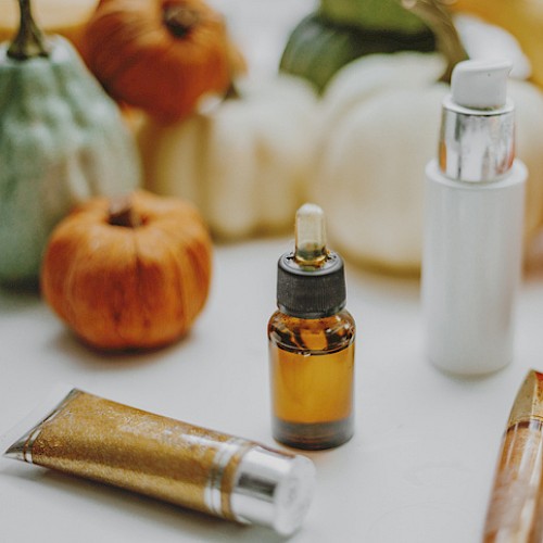 This image shows various small pumpkins, skincare products including a dropper bottle, a pump bottle, a tube, and a lipstick, placed on a white surface.