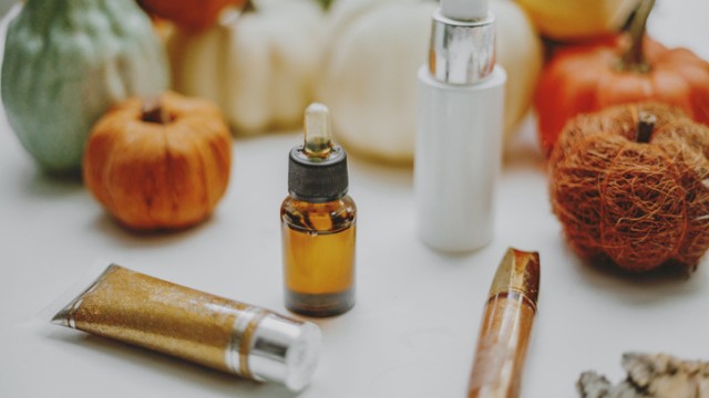 This image shows various small pumpkins, skincare products including a dropper bottle, a pump bottle, a tube, and a lipstick, placed on a white surface.