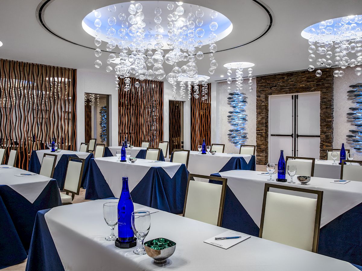 A modern conference room setup with white tablecloths, blue glass bottles, and elegant lighting decorations hanging from the ceiling.