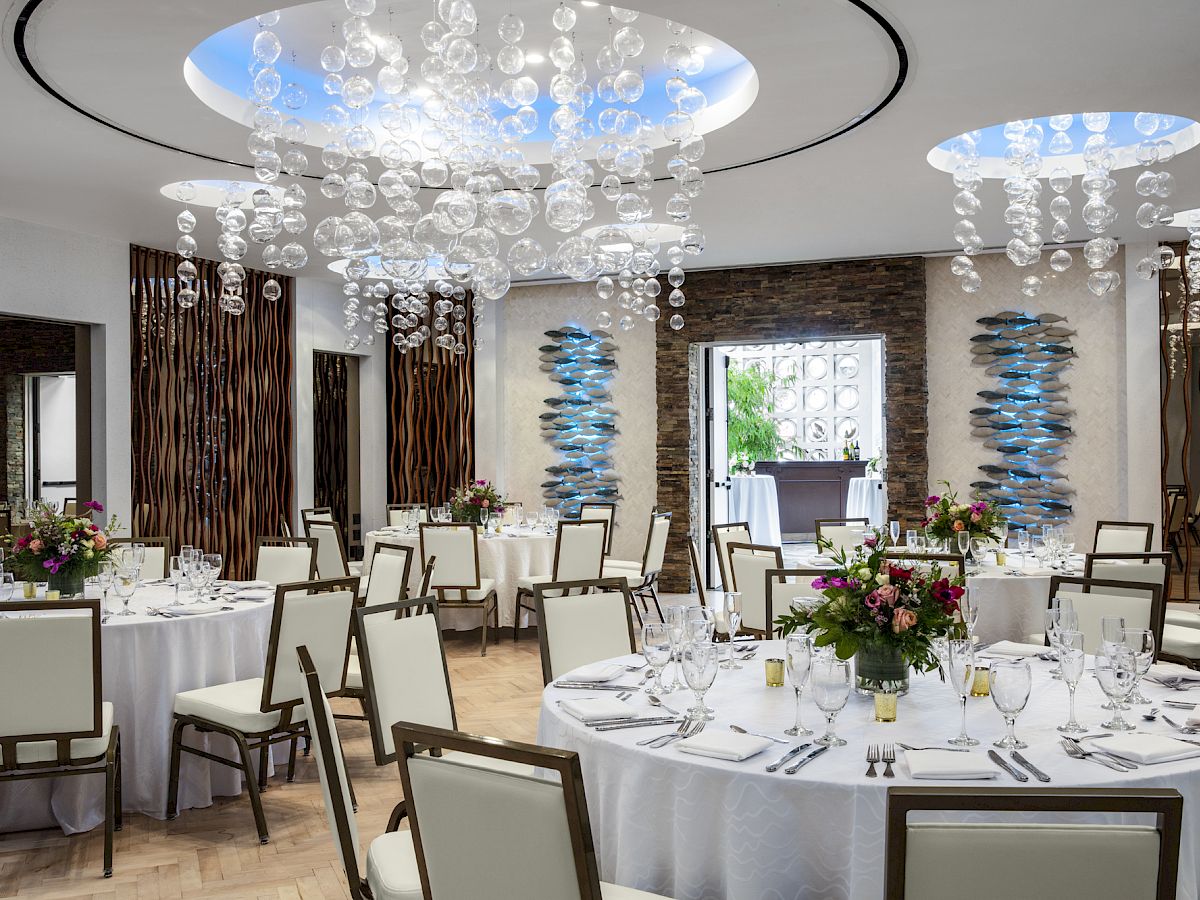 The image shows an elegantly decorated banquet hall with round tables set for an event and modern chandeliers hanging from the ceiling.