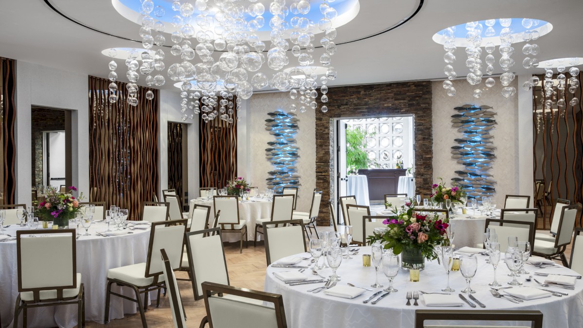 The image shows an elegantly decorated banquet hall with round tables set for an event and modern chandeliers hanging from the ceiling.