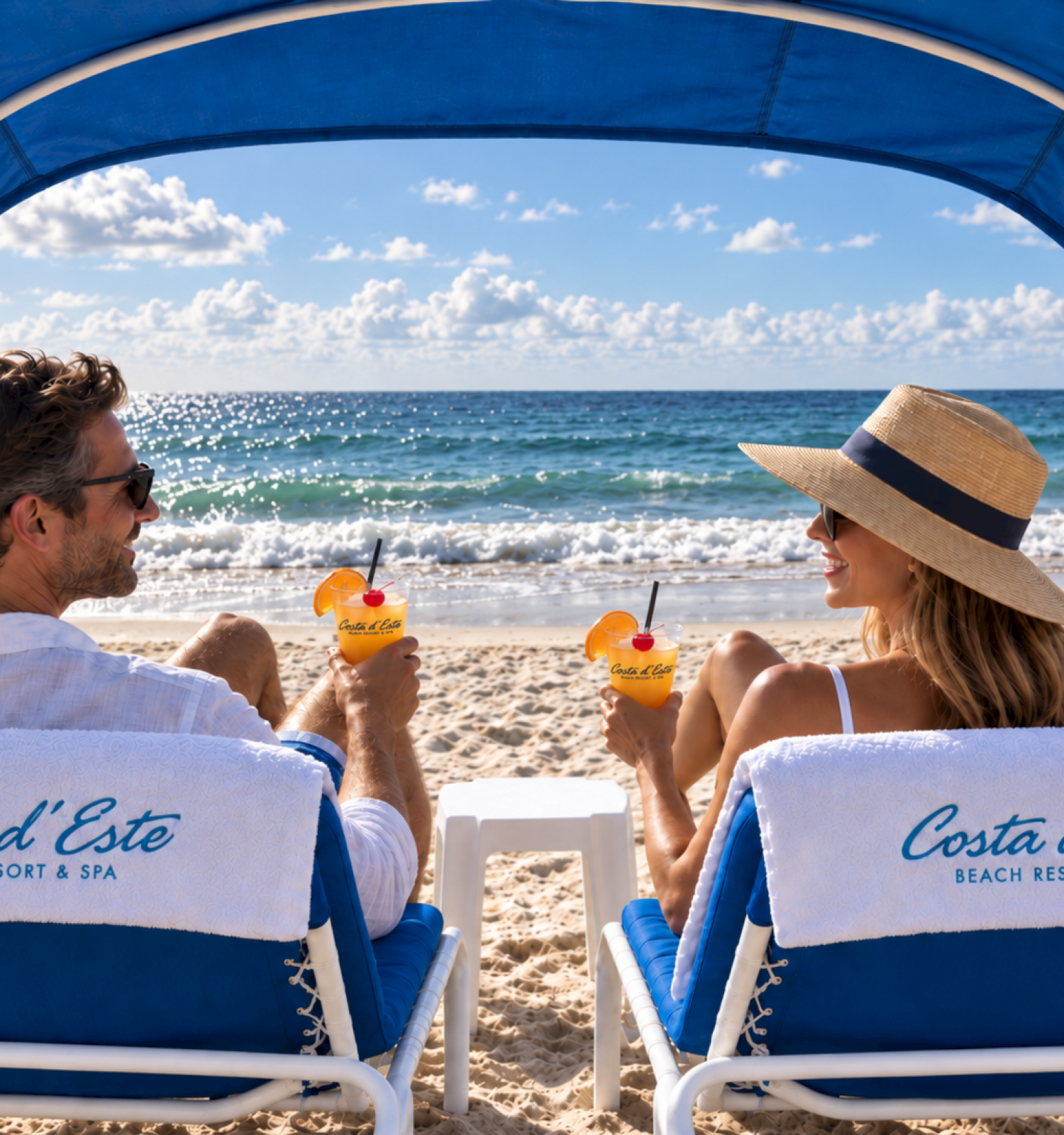 A couple relaxes on beach chairs, enjoying drinks under a canopy with the ocean and sky in the background.