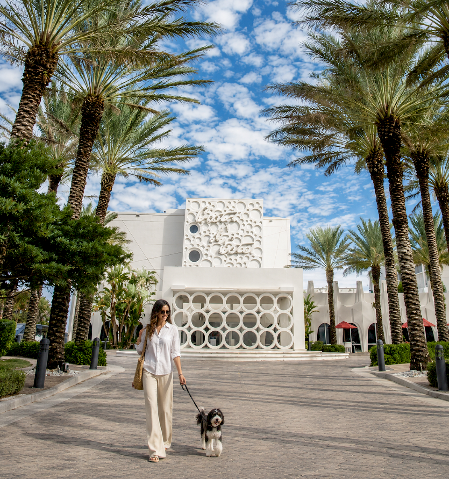 A woman walks her dog in front of a white building with circular designs, surrounded by palm trees on a sunny day with a blue sky.